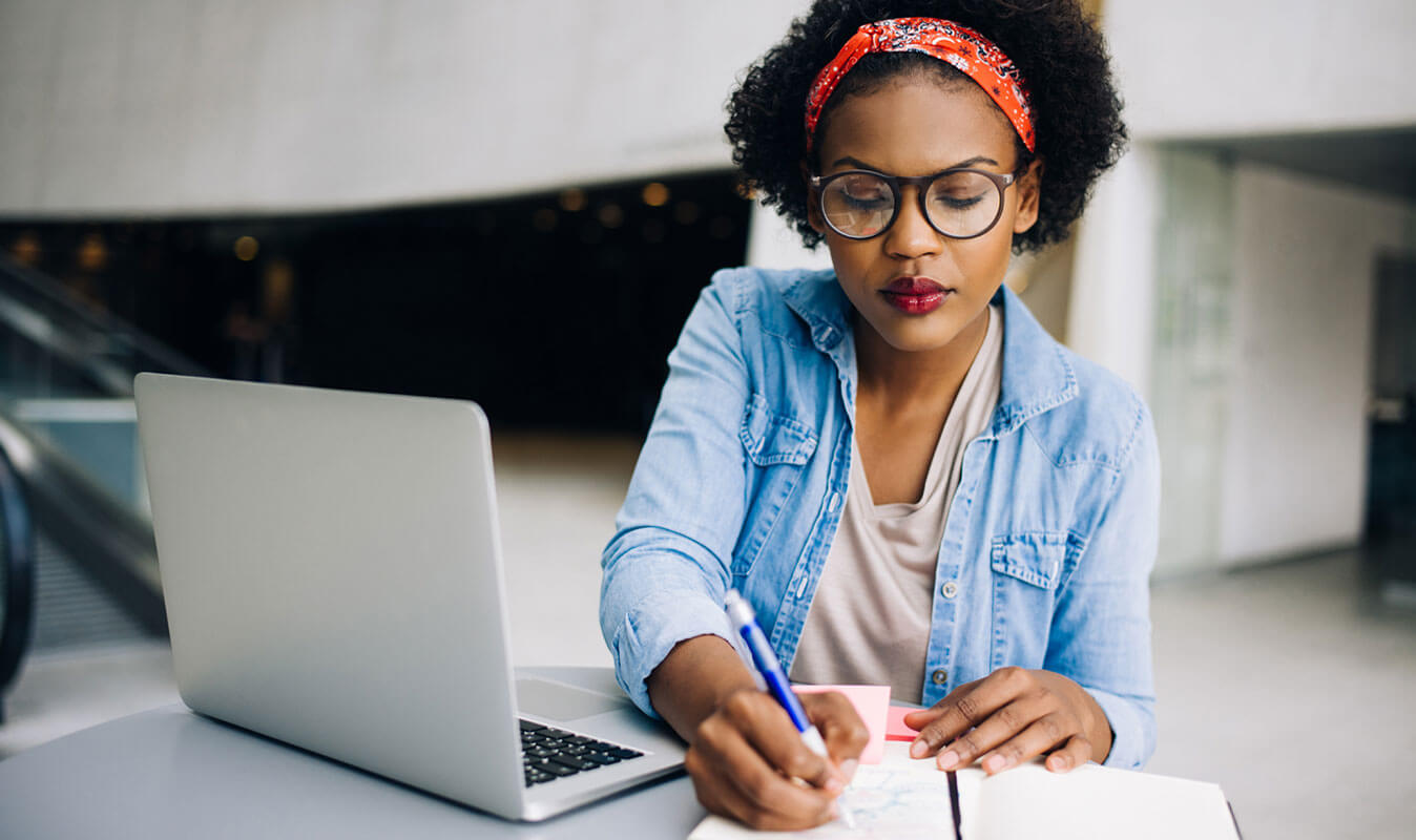 A woman writing something on a notepad whilst working on a laptop